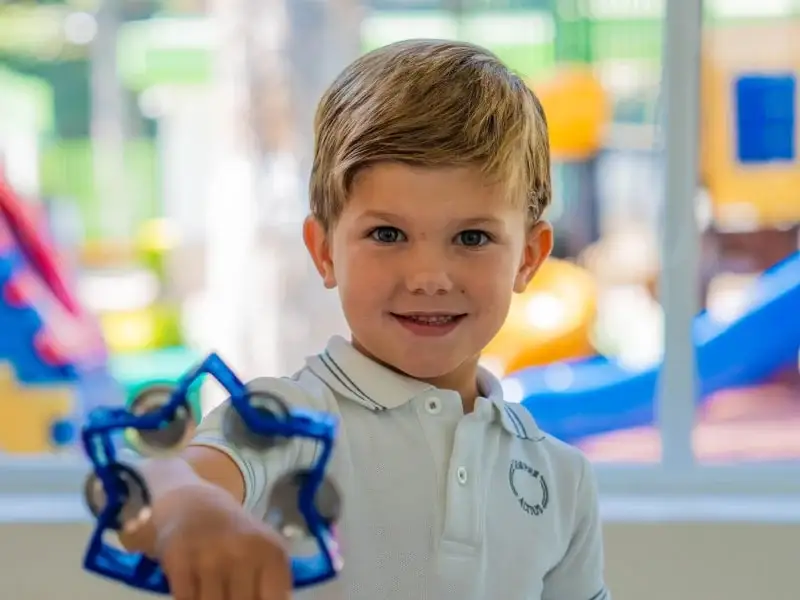 Niño sostiene juguete educativo en aula con parque infantil de fondo en escuela