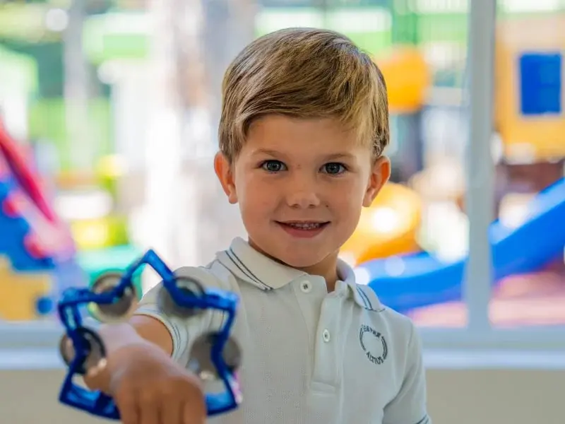 Niño sostiene juguete educativo en aula con parque infantil de fondo en escuela